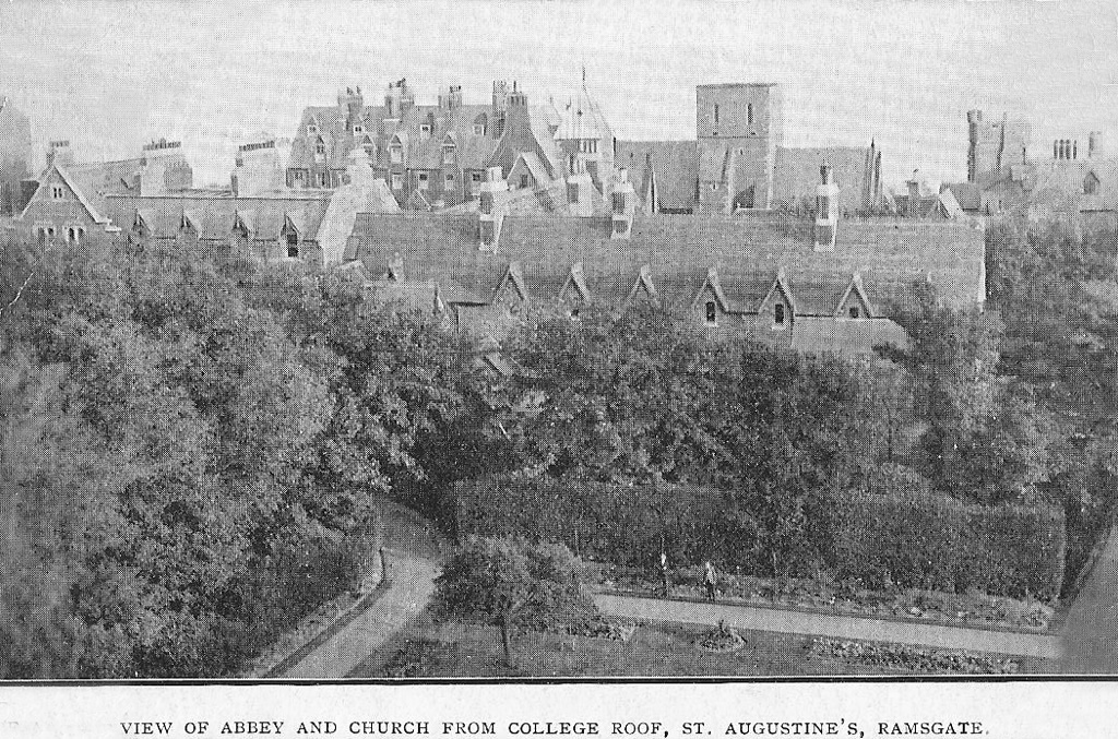 greyscale photo\' of long multi-storey buildings with dormer windows, and a high square church tower beyond, all partially screened by trees in the foreground