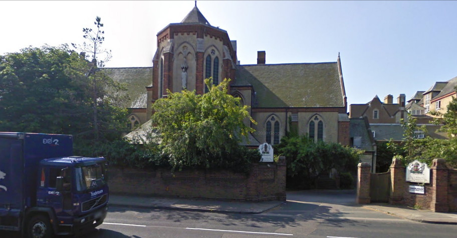 church of creamy-beige brick trimmed with red, with a short round tower sticking out on the side nearest the viewer and more buildings to the right, seen behind bushes and a low wall