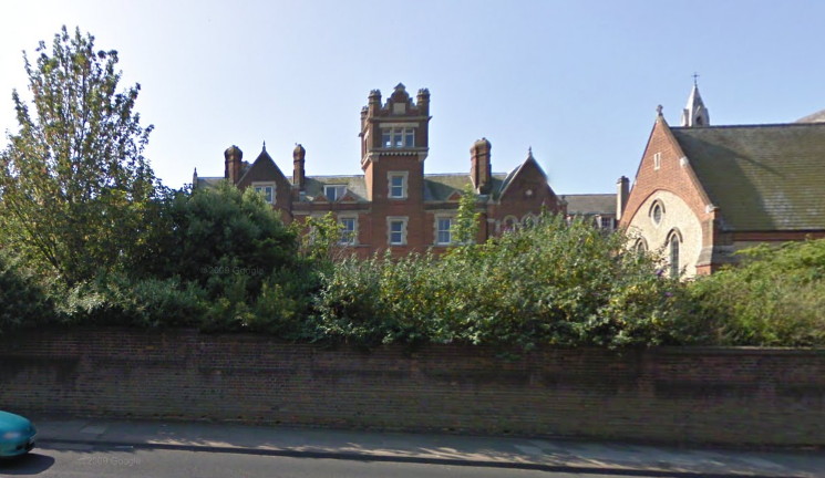 large red-and-cream brick building with a central tower with a crenellated top, glimpsed behind bushes and a low wall, with other buildings to either side