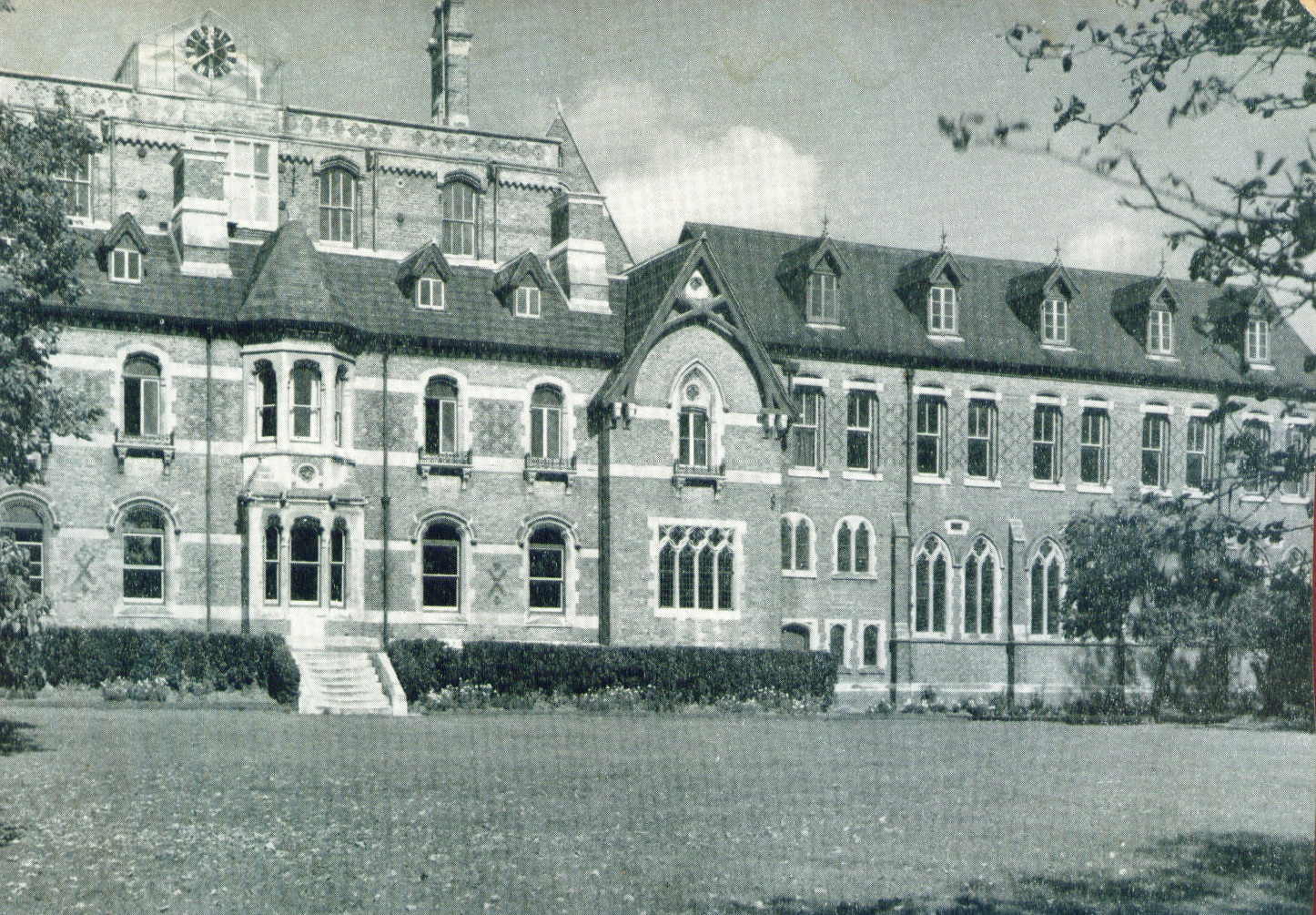 greyscale image of a medium-sized Victorian brick house with cream banding, with a two-storey bay window and flight of white steps at centre and a gable-end to the right, with a three-storey extension to the right of it, bigger than the house itself, and a four-storey extension behind it, looming over the roof