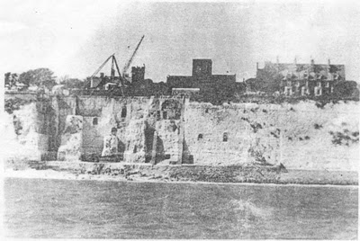 faded greyscale photo\' looking from the sea towards a high white cliff, on top of which can be glimpsed an elaborate house and a large church with a square tower