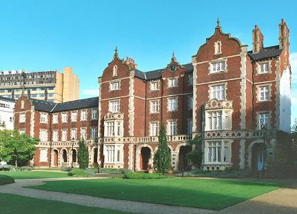 Victorian building in red-brown brick with white trim, with a central four-storey front with three arches at bottom leading to recessed windows and doors, flanked by square projecting blocks the lowest two floors of which have ornate bay windows and which have curvy roof-lines: on the left is a long wing with more arches along the bottom and ending with another projecting square block while the right side comes to a blank end: all seen diagonally across a flat lawn with modern office blocks visible beyond