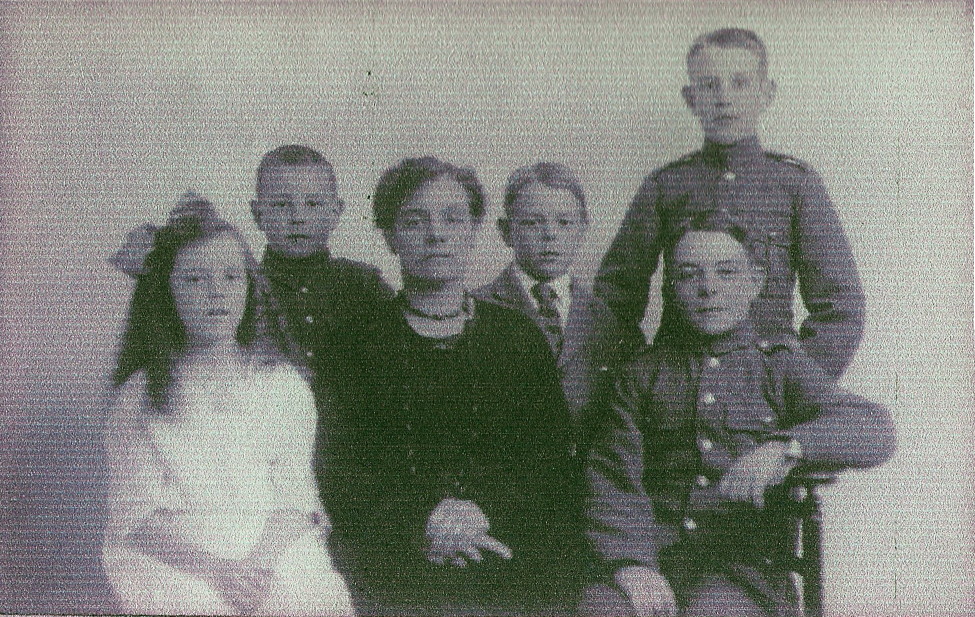 black and white photo\' showing a woman with an oblong face, short or pinned-up dark curly hair, a dark dress and a choker necklace sitting surrounded by a little girl with long ringlets, a huge bow in her hair and a white dress and four boys of varying ages, one in school uniform and the others in army uniforms