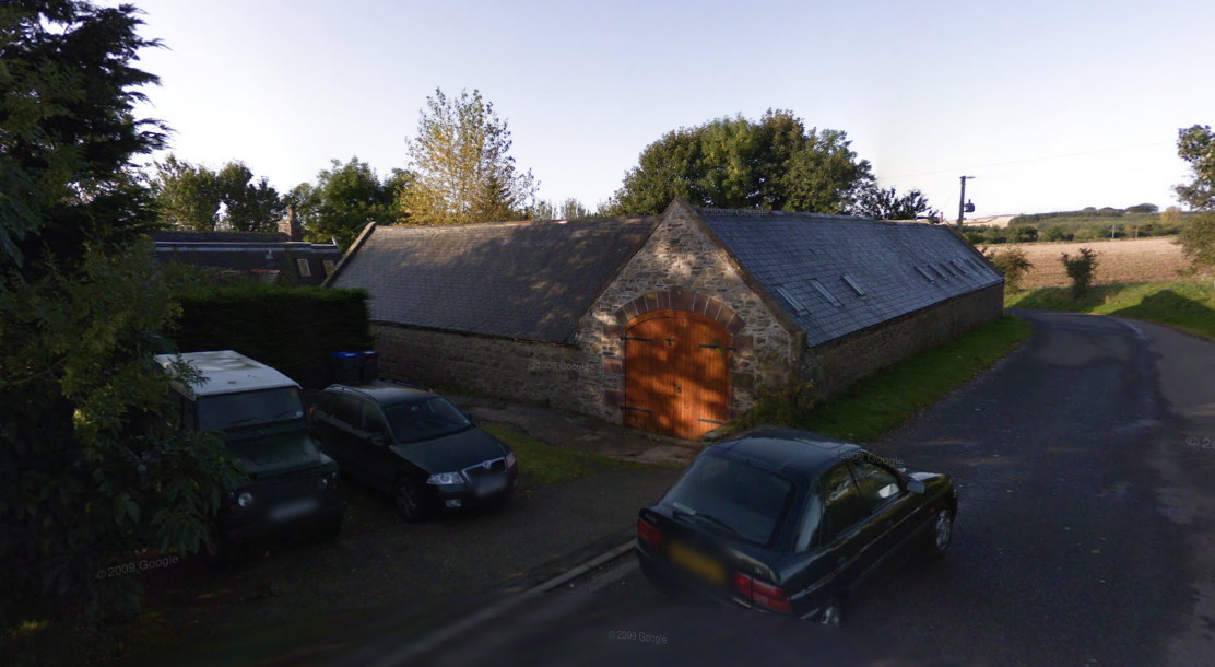 view crosswise across a road towards a group of low dark grey farm buildings, especially a long building with blank walls, a huge wooden door in the end face and windows in the roof; there are cars around the buildings