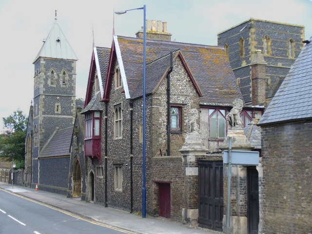 view diagonally along a road edged first with a complex Gothic building in grey-brown brick, then a church in slate-grey