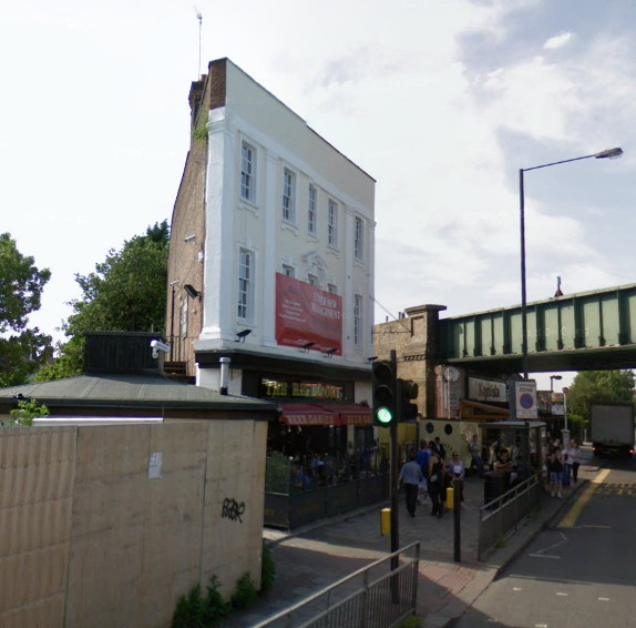 almost triangular buiding, seen from one of the corners, with a high white front with dolls\'-house-style windows, with a dark pub frontage at the bottom, trees behind the building to the left and a railway bridge crossing the road at the right