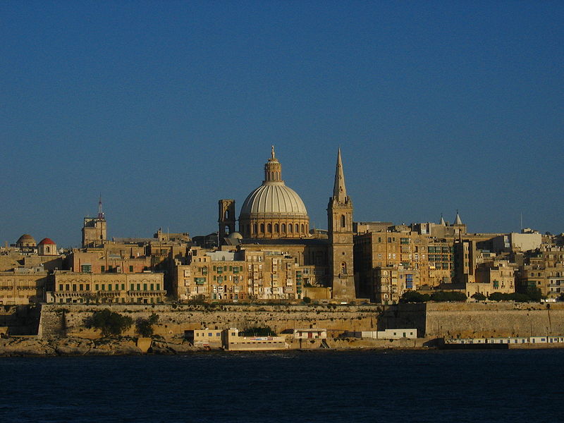 view across water under an overcast sky towards a jumbled cityscape of old and newish buildings, from a great domed church down to slabs of 1960s-style blocks of flats, all in pale snuff-coloured stone