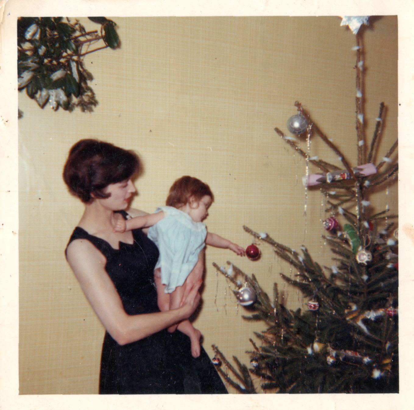 lean woman in a dark blue dress, with brown hair in a medium-length \'60s cut, holding a baby who is reaching out to touch a bauble on a Christmas tree