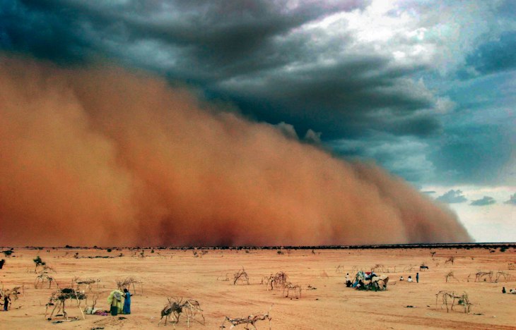 billowing cloud of peach-coloured dust rolling in across a sandy desert, watched by tiny human figures standing beside makeshift shelters