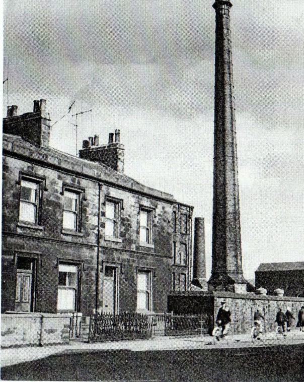 black and white photograph of small square terraced houses with towering factory chimney in background