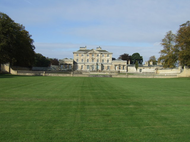 view across a flat smooth lawn to a large pale house with side-wings, and a low wall in front of it