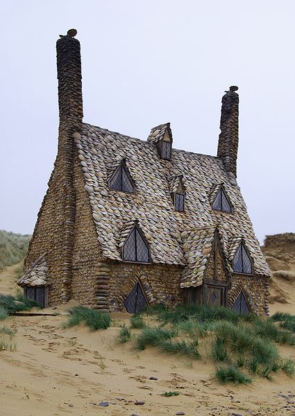 three-storey cottage covered in brown shells, standing on a beach, with very high, steeply-pitched roof with two rows of dormer windows, one above the other