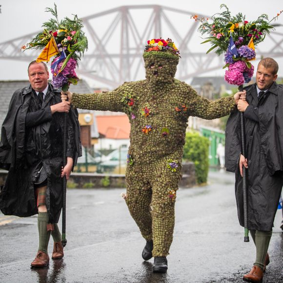 Photo of a figure covered in burrs and holding out two staves topped with flowers at arm\'s length, with a man on either side helping to support the staves