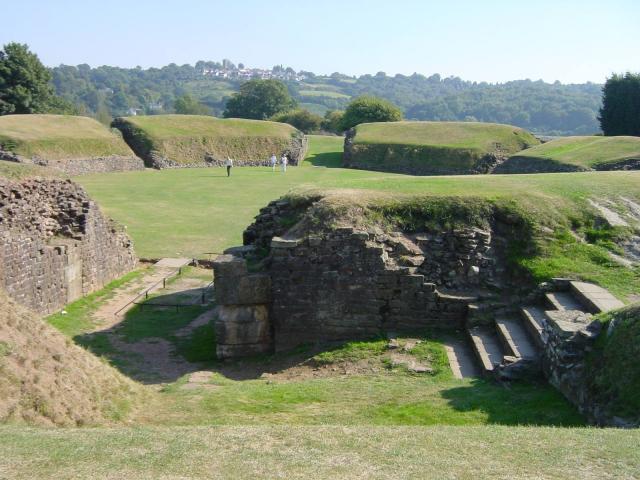 view past steps and broken stone walls, through an entrance into a green arena surrounded by pillow-like, grassy mounds roughly in the shape of blocks of seats rising in tiers, with walkways in between
