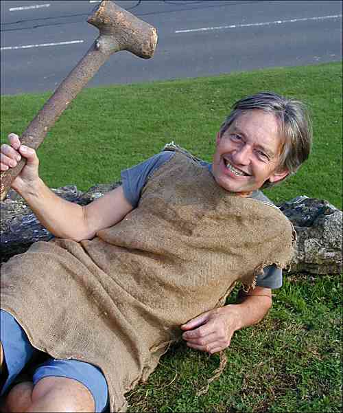 John at 62 wearing a sackcloth tunic over a light blue shirt and shorts, sprawled on his back on grass clutching a hammer-shaped chunk of branch and grinning like a maniac