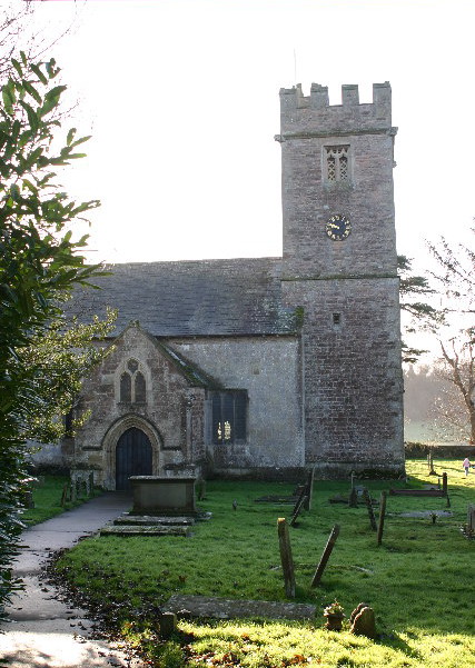 ancient church with a large porch facing the viewer and a square tower to the right, with a black and gold clock on the tower, ancient, leaning graves around the church and a grassing slope glimpsed rising up towards a wood behind the church
