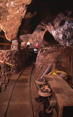photo\' of an underground cave, with jagged red-brown rocks, some hanging down from the ceiling, and a railway track between low brick walls, arcing across the cave and disappearing into a tunnel: beside the track in the right foreground a small wooden goods-wagon lies on its side, and in the distance a group of tourists stand on a bridge overlooking the track