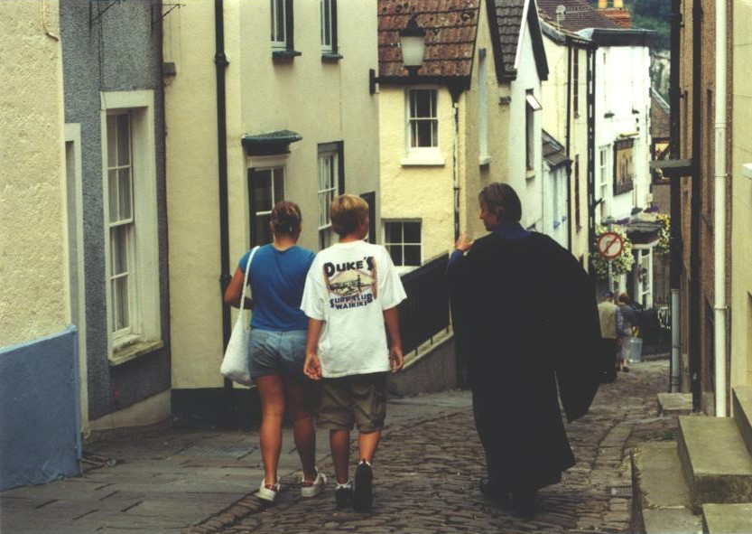 very steep, winding cobbled street with man in robes and two people in hiking gear walking away down it