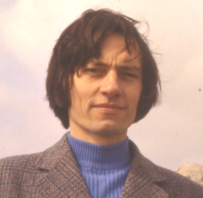 full-face head and shoulders shot of John looking very handsome, outdoors with his hair blowing across his face, wearing a hyacinth-blue polo-neck sweater and a brown tweed jacket