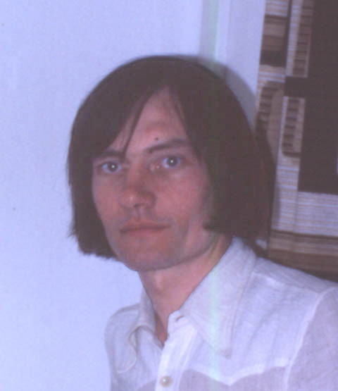 head and shoulders shot of John looking very handsome and poetic, standing with his left shoulder towards the viewer and his head turned to look at the camera, wearing a white cheesecloth shirt and with his long black hair neatly arranged just below the jaw, standing in front of a white wall and a hideous brown and white 1970s curtain