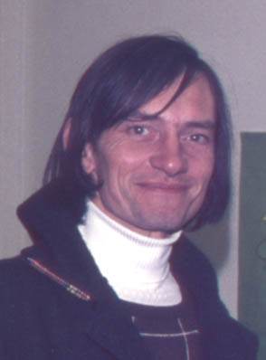 head and shoulders photograph of John, seen slightly from his right but looking at the camera, smiling, wearing a high white turtle-necked sweater, something odd like a dark tartan waistcoat only not quite, and a thick woolly sort of black coat or jacket with a high Dracula collar