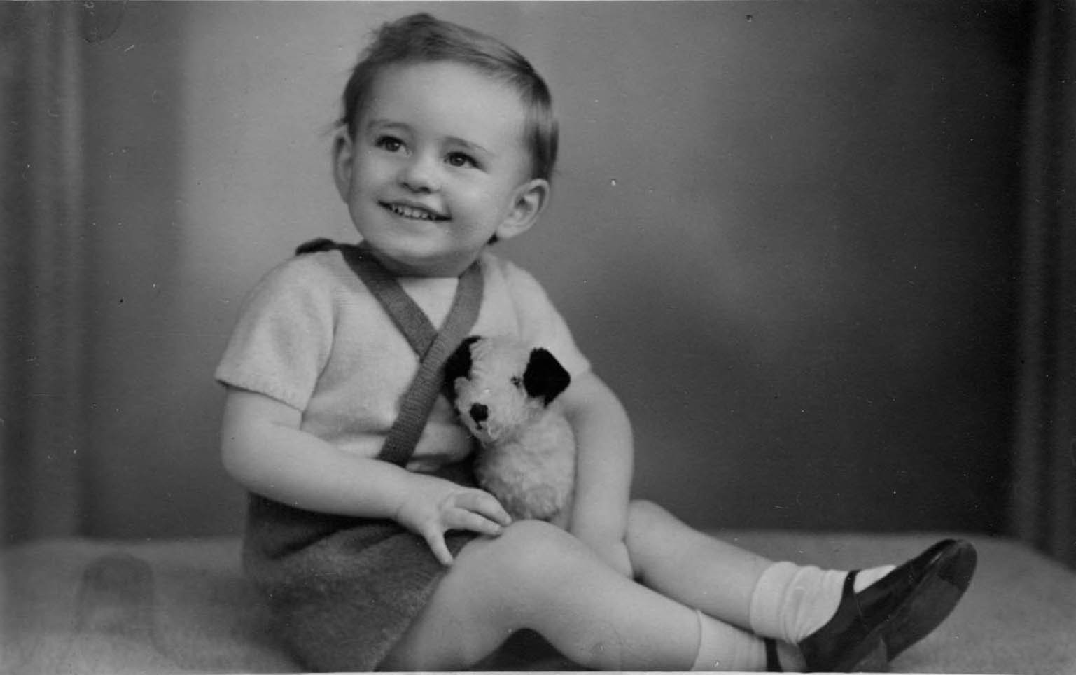 John as an extremely pretty toddler, sitting on the floor, smiling broadly and holding a black and white toy dog, and wearing a white shirt, dark shorts with braces crossed across his chest and black patent shoes with white socks