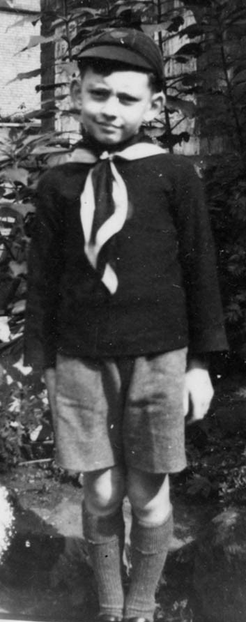 black and white shot of a young boy, standingly rigidly as if \'at attention\' in front of some bushes, wearing shorts, a thick dark shirt and a dark school cap, with a dark and white scarf tied round his neck
