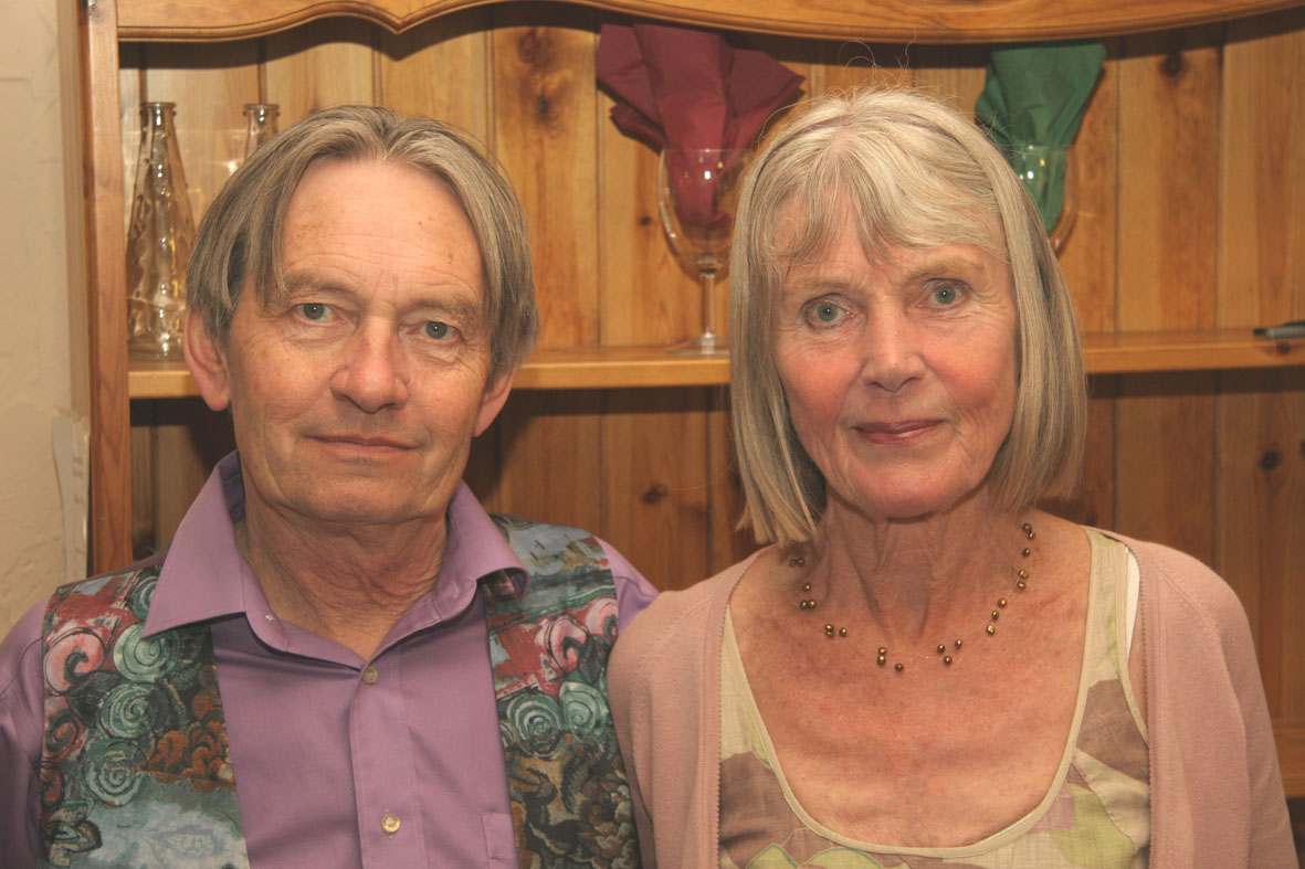 head and shoulders shot of John and Shirley side by side, facing the camera, both grey-haired, both looking pleasant and fond, John wearing a soft dull purple shirt and blue, grey and terracotta swirly waistcoat, Shirley in a dusky pink cardie over a dressed patched with a similar pink on white