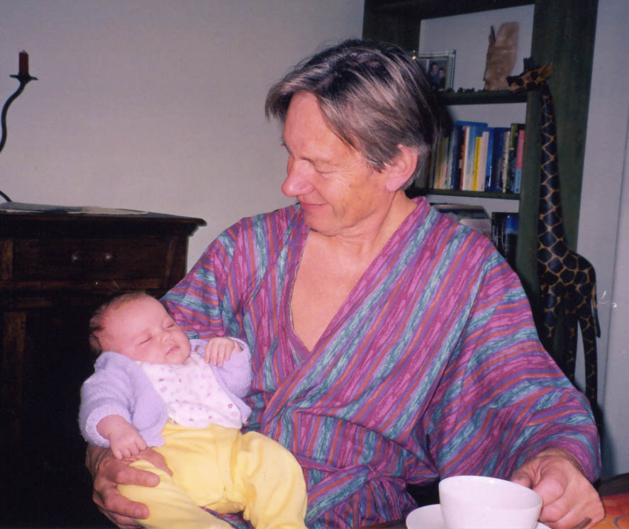 John in his sixties sitting wearing a loose, lightweight dressing-gown striped with smudgy longitudinal stripes in light blue and mulberry over a bare chest, clutching a tea-cup in his left hand and with a small baby dressed in a white top and lemon leggings folded into the crook of his right arm, gazing down soppily at the baby