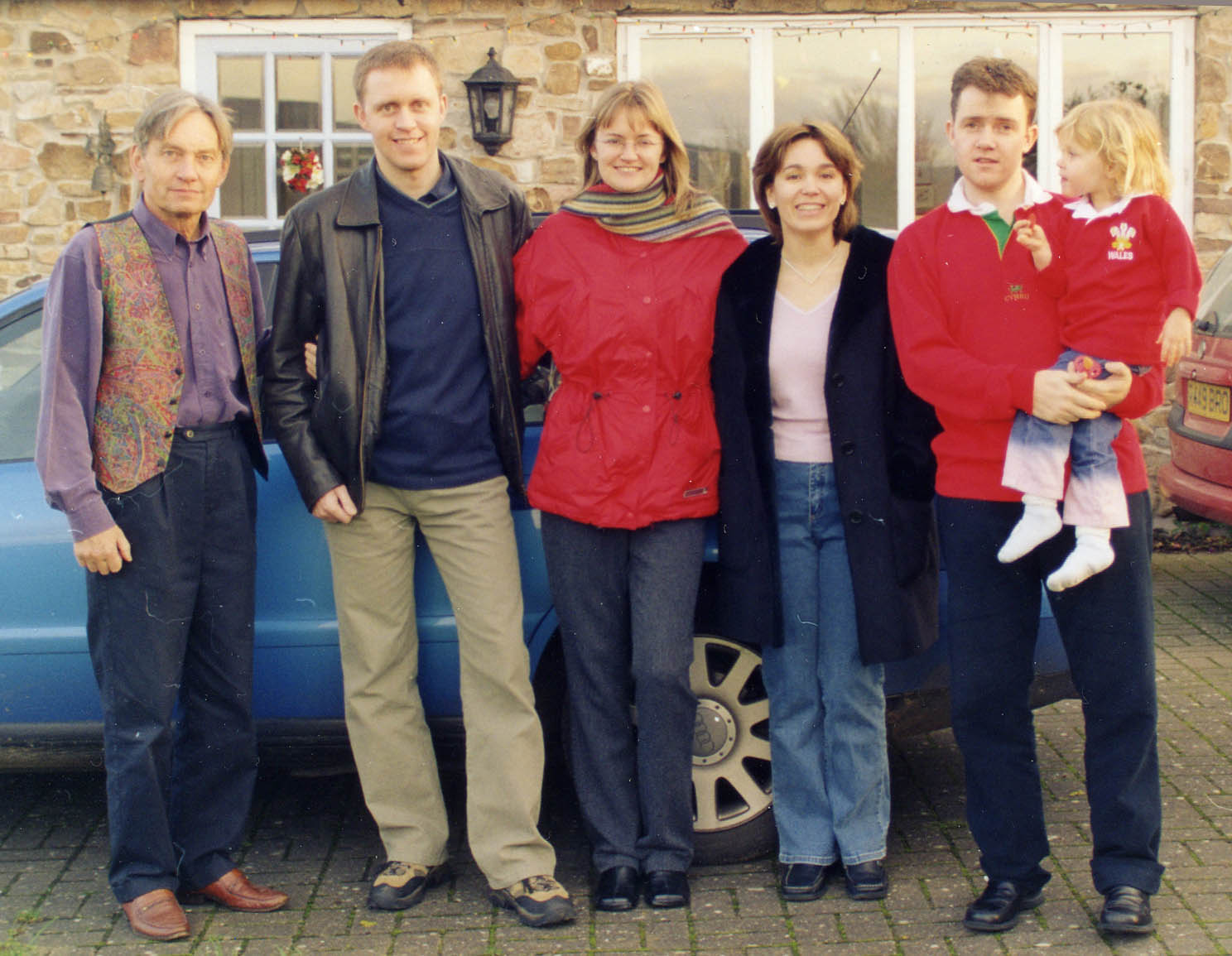 a row of people standing in front of a blue car in front of a stone wall and windows: John at far left, wearing jeans, a purple shirt and autumnal waistcoat; then a young man with John\'s features but nearly a head taller; two young women and another young man holding a blonde child about four years old