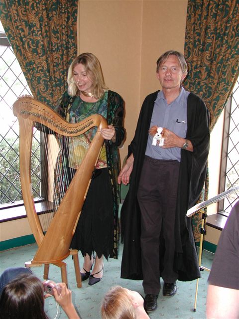 photograph of a young, blonde woman in floaty clothes playing a very large harp; John is on her left singing, wearing a black academic gown over dark purple trousers and a light blue shirt and holding a Beany Baby ginger and white spaniel in his left hand; they are standing in the corner of a room between two large windows with drapes