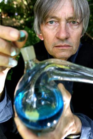 John at 67, wearing a black academic gown, gazing piercingly at the camera across the top of an alembic from which he has just removed the stopper