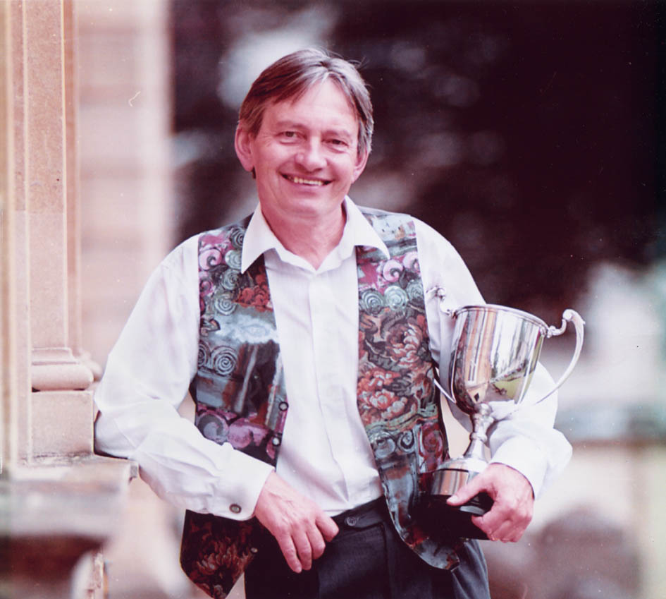 John in a swirly blue, grey and terracotta waistcoat over a white shirt, lounging leaning his right elbow against the architecture, holding a big silver cup in the crook of his left arm and grinning