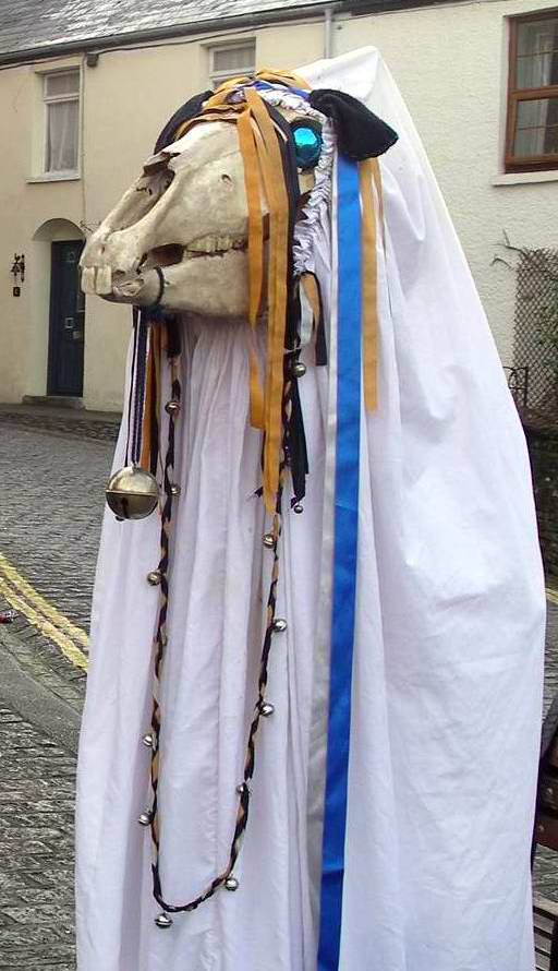 photograph showing a horse\'s skull, draped with ribbons and bells and with leather ears and gleaming blue-glass eyes, draped from the ears back in a long white cloth underneath which someone is holding it aloft, against a backdrop of a street of small terraced early 19th C houses