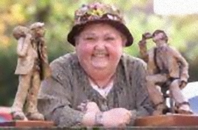 photo\' showing a round-faced, jolly, grinning elderly woman in a grey cardigan and a floppy black hat with flowers round the brim, leaning her elbows on a bench on which are two pottery figures of workmen
