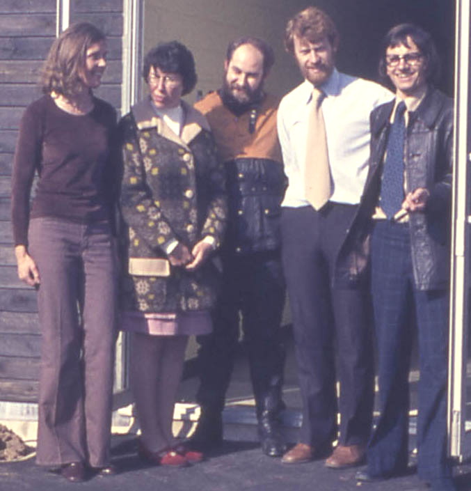 5 people standing in front of a brown wooden building: from left to right a tall slim woman with longish brown hair, in dull purple slacks and sweater; a woman of medium height with black hair, in carpet-like hippy coat; a shortish, hefty bearded bloke in a navy shell suit with orange upper chest and shoulders; a tall bearded bloke with a mop of light brown hair, in jeans and a white shirt and apricot tie; and a slight bloke with long black hair and glasses, in jeans, a leather jacket and a blue kipper tie