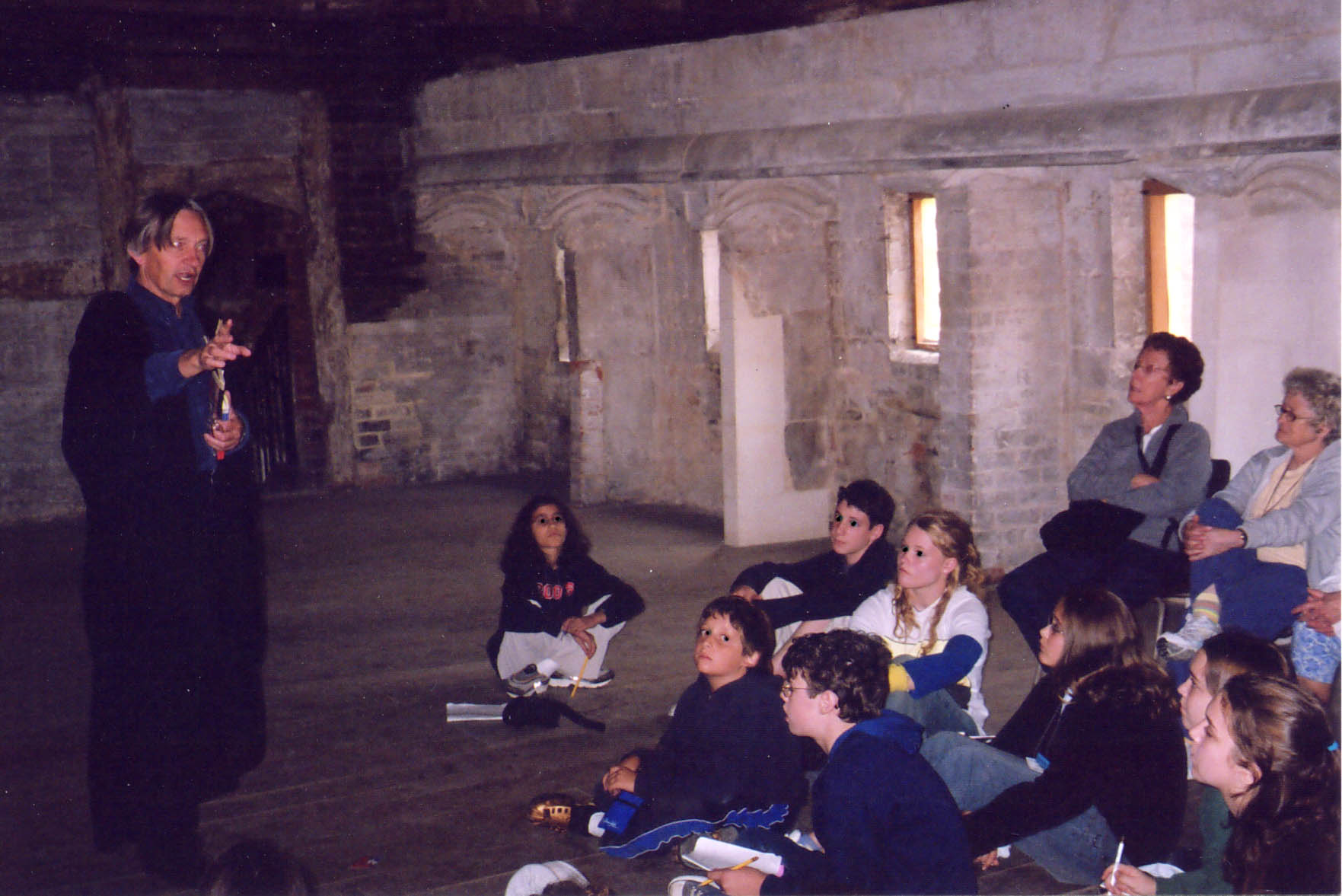 John standing in black academic gown gesturing and declaiming to a group of children seated on the ground in a hall which forms part of an ancient monastery, edged with battered stone buttresses which have been partially restored with plaster