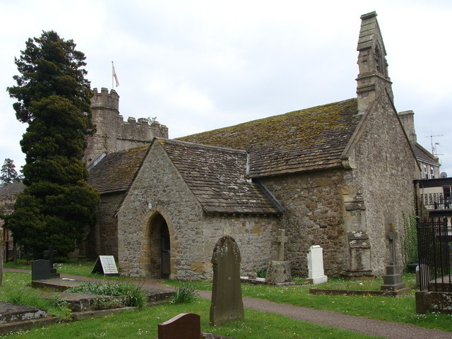 small grey-brown Mediaeval church with a large plain porch, seen diagonally from the right,  with an ornate spike of carved stone sticking up from the gable at the near end, and a split-level, crenellated square tower at the far end half-hidden by a large tree, and with old gravestones around the building