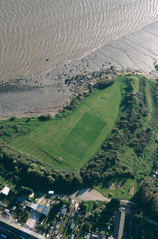 aerial view looking down and across at an angle at one corner of a triangle of grass bounded on the near two sides by an enormous earthwork: at the top of the picture a cliff edge falls away down to a silted beach and on the right the ground slopes down sharply below the earthwork and is striped with terraces