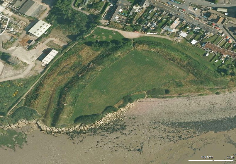aerial view showing a strip of modern buildings at the top and a strip of silted beach at the bottom, with between them a large triangle of grass on which is one corner of an enormous, roughly square earthwork the rest of which has been washed away
