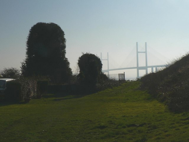 grassy track at evening, with an earth bank on the right and on the left two tall stone stumps covered in ivy, with a suspension bridge glimped in the distance