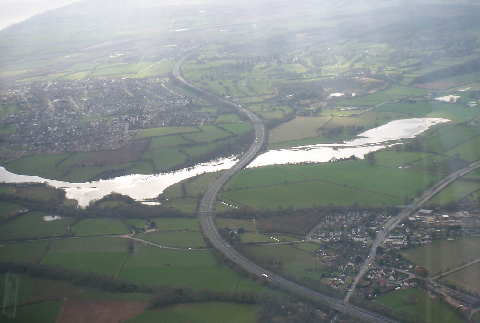 aerial view of a long thin lake with irregular edges, shining like quicksilver, unrolled from right to left across a flattish landscape of fields in between a large and a small village, with a motorway snaking across the landscape and crossing the lake by bridge