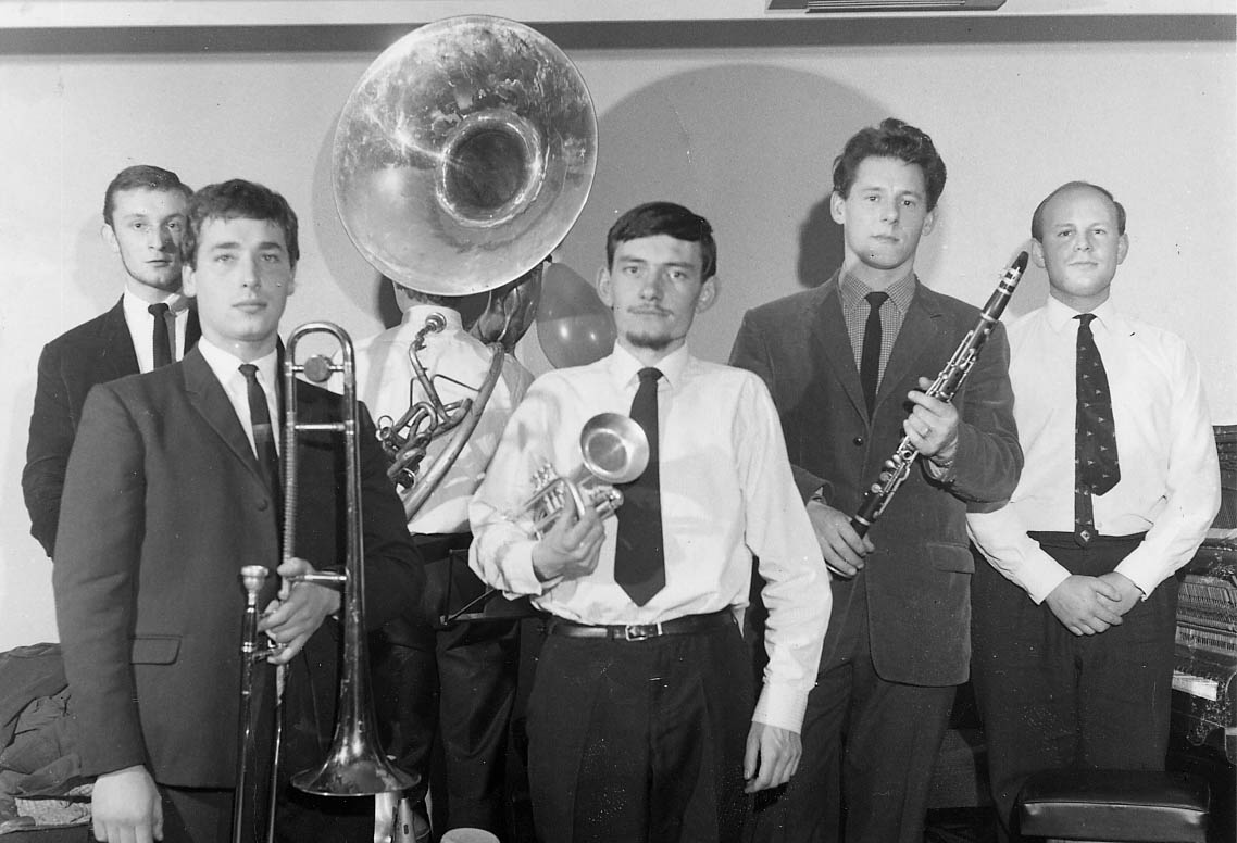 six young men in 1960-ish clothes stand facing the camera (except the tuba player who is facing away) and holding various musical instruments: John is at centre front with a cornet, white shirt and dark tie, with a thin moustache and a beard which is just dark fluff under his chin, and his hair combed horizontally across his forehead from a low parting