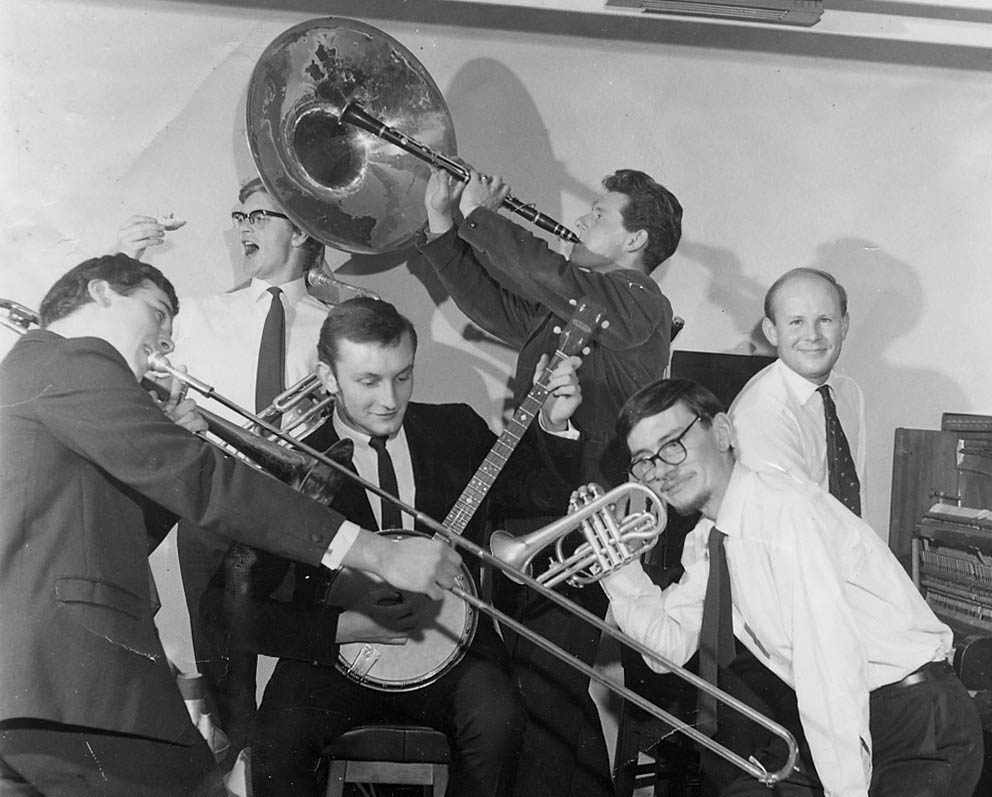 six young men in 1960-ish clothes playing various musical instruments: John is at right front with a cornet, white shirt, dark tie and dark-framed spectacles, with a thin moustache and a beard which is just dark fluff under his chin, and his hair combed horizontally across his forehead from a low parting