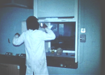 back view of a man in a white lab. coat, with long black hair, holding some small object aloft in front of a fume cupboard