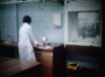 back view of a man in a white lab. coat, with long black hair, working at a laboratory bench next to a fume cupboard