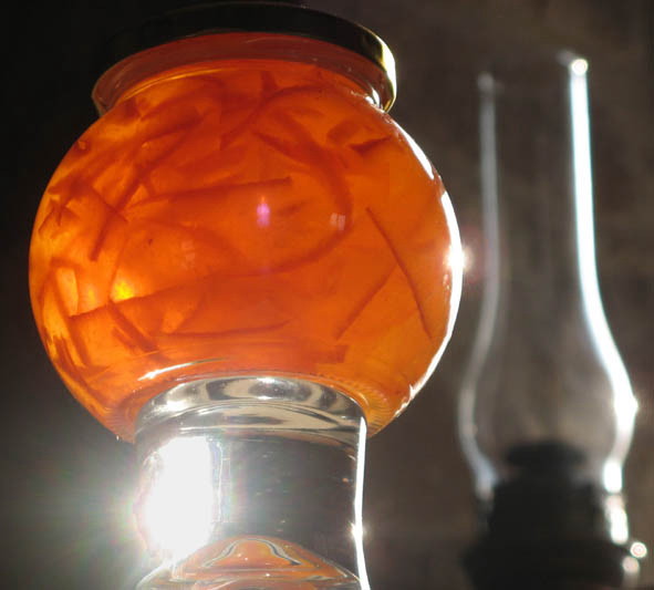 light glowing orange through an almost spherical jar of marmalade, balanced on a drinking glass which forms a clear plinth, set slightly left of centre with a glass gas-lamp in the right background