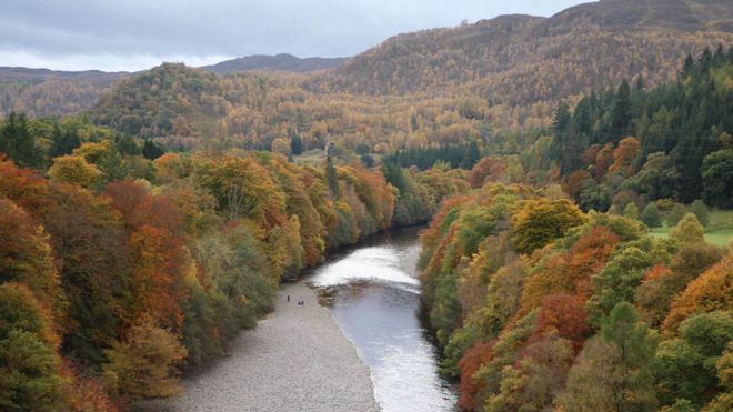 view from a high place along a small river flanked by trees in autumnal colours, with wooded hills in the distance