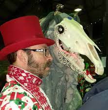 Photo of a man in a red top hat leading a figure with a horse\'s skull for a head