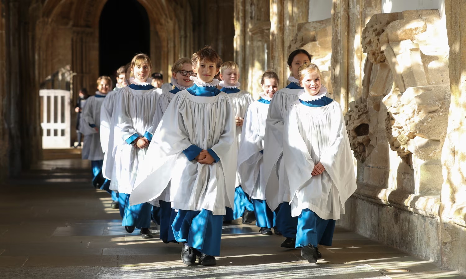 Photo of several boys wearing blue-collared white surplices over blue trousers, striding along an aisle past a carved stone wall